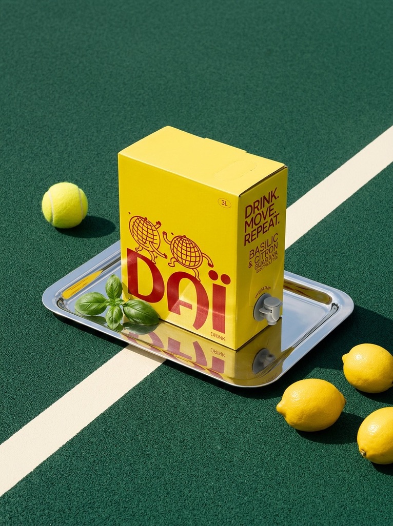 Une fontaine à boisson Daï jaune sur un plateau argenté, posée sur un court de tennis vert, entre citrons et balle de tennis.